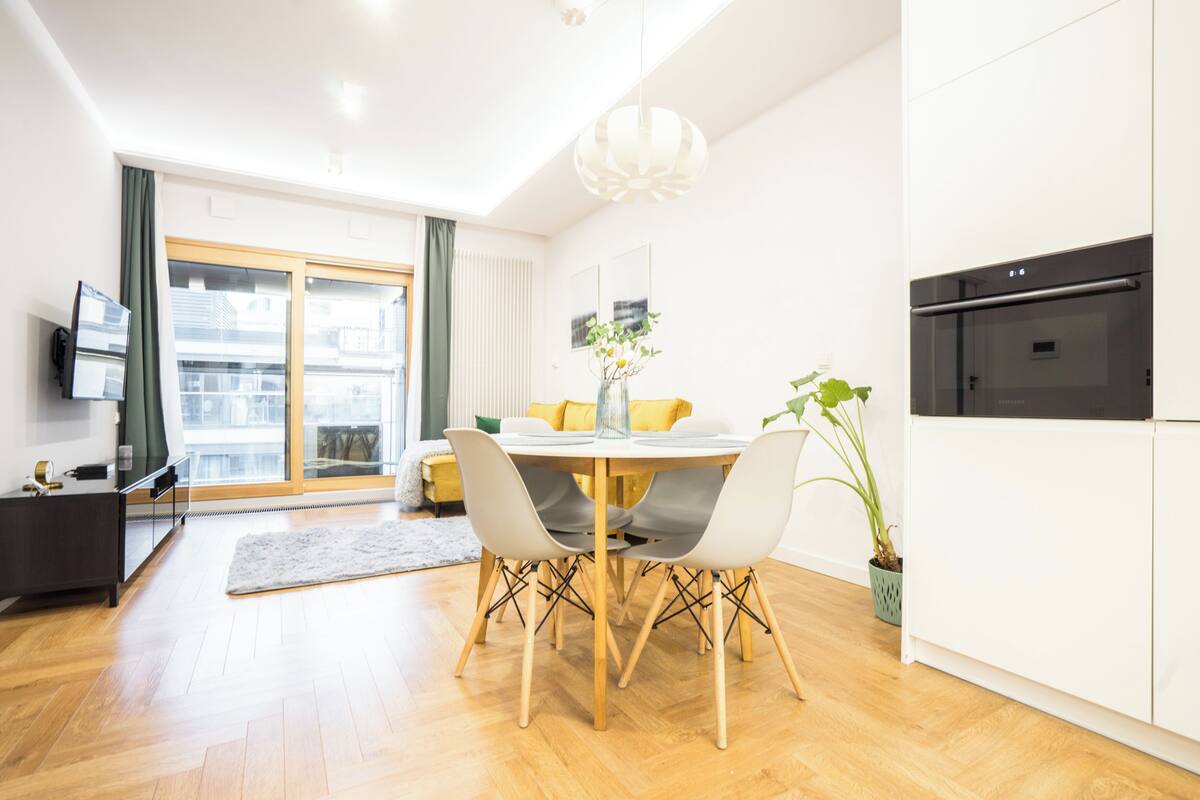 A modern living room with a dining area, a kitchen with a built-in oven, and a large sliding glass door leading to a balcony.