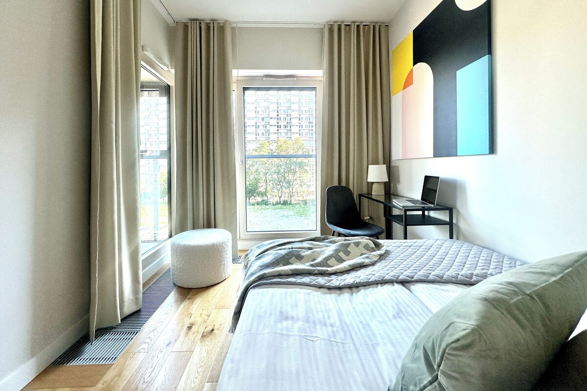 A modern hotel room with a large bed, a desk with a laptop, a chair, and a view of the city through the window.