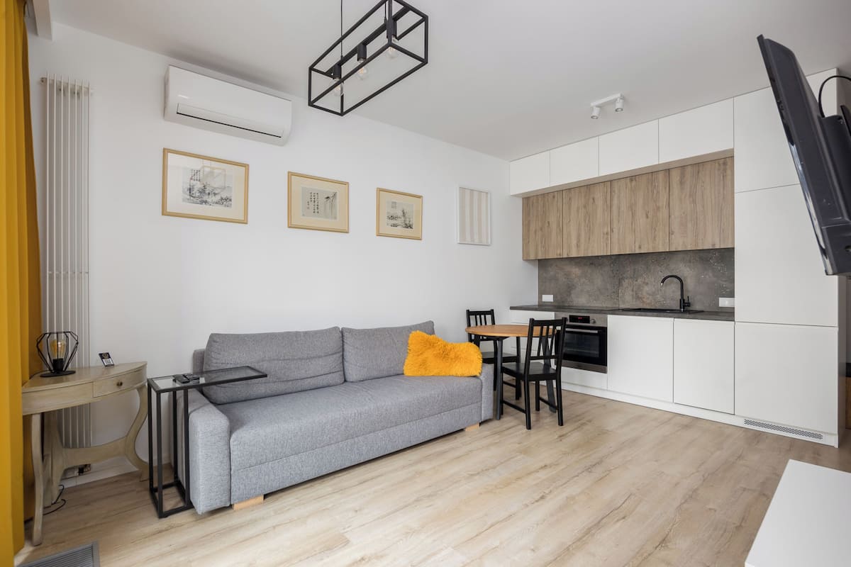 A modern living room with a grey sofa, a small wooden dining table, and a kitchen area with white cabinets and a wooden countertop.