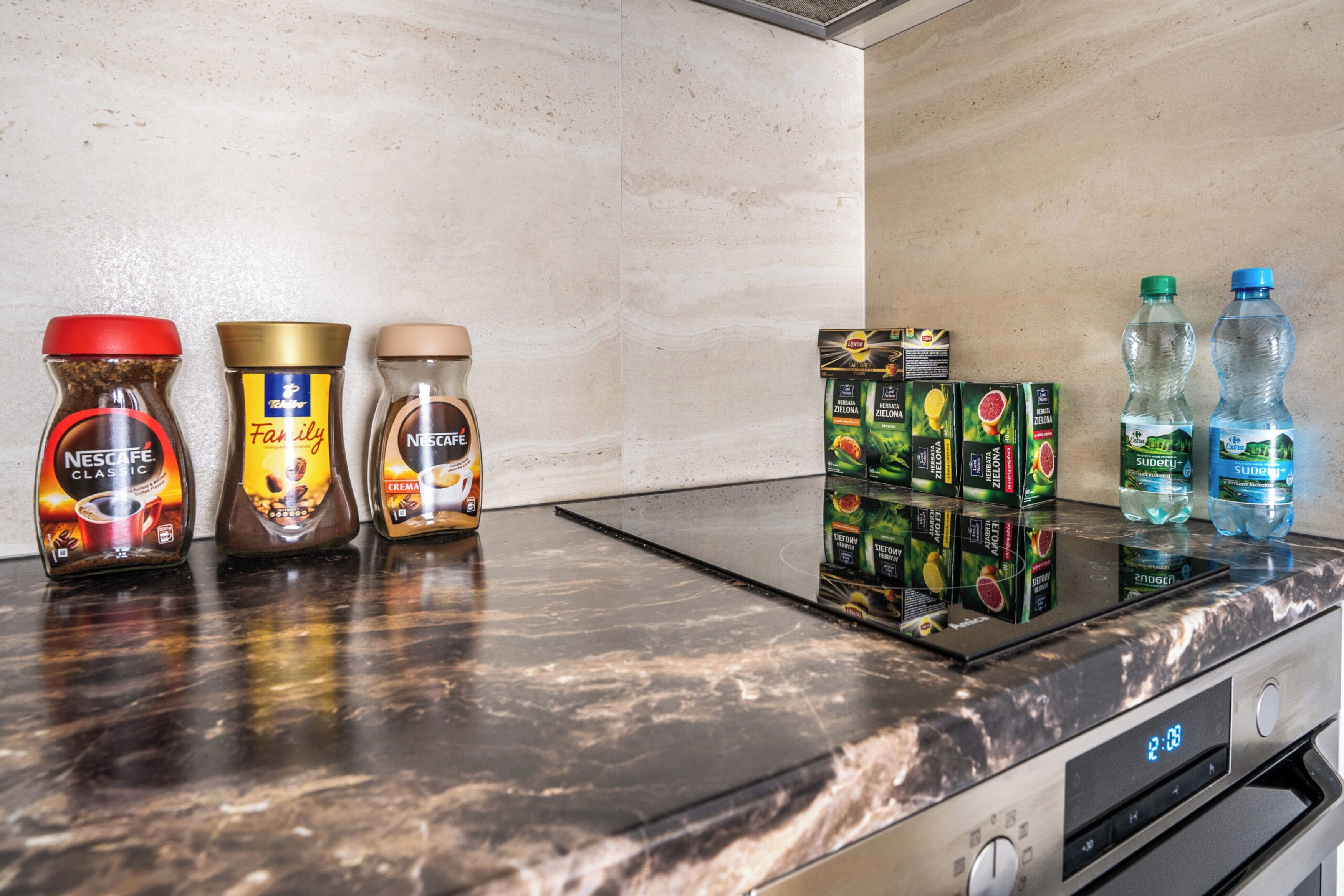 A kitchen counter with Nescafé Classic, Family, and Nescafé Crema coffee jars, a box of Supercafé Zelnova, and two water bottles.