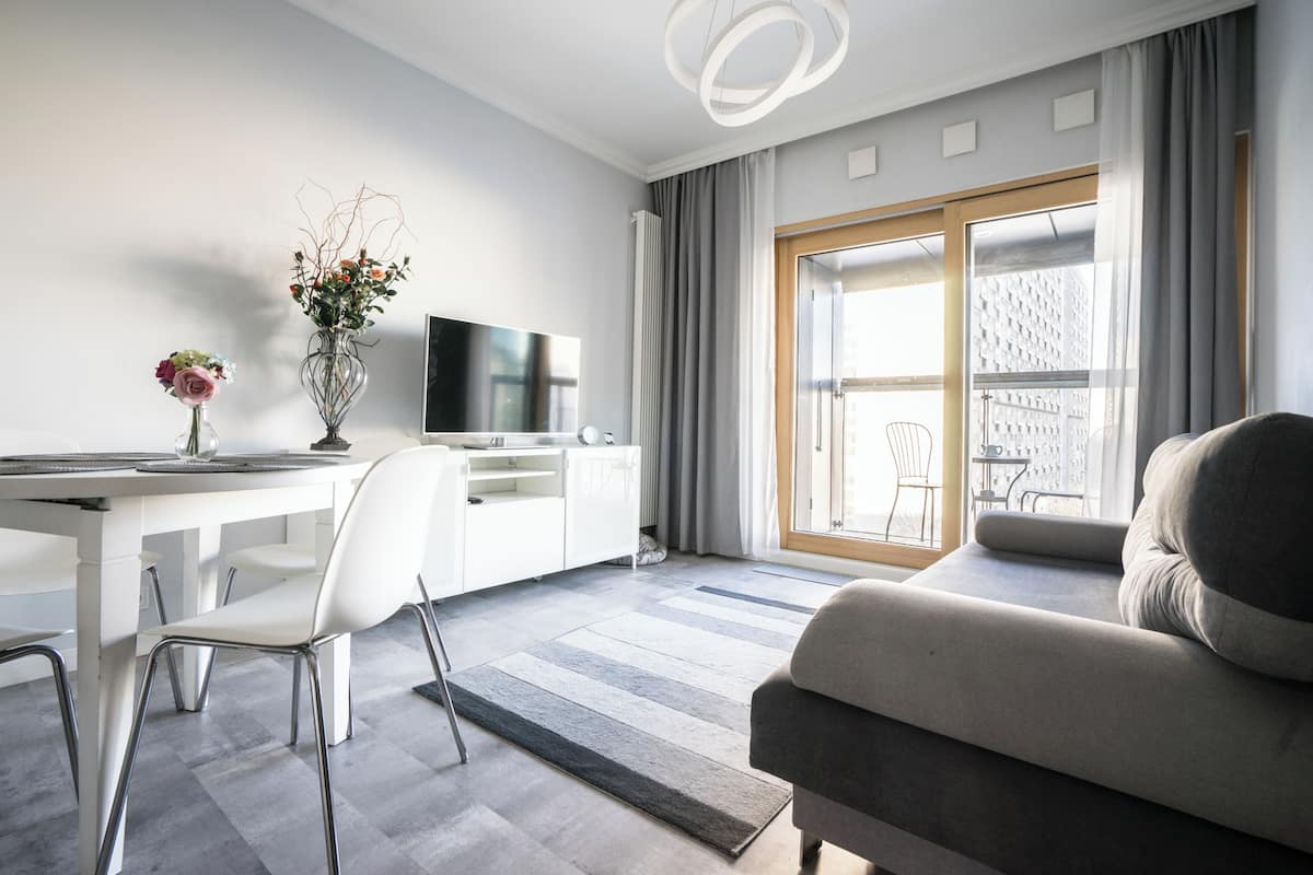 A modern living room with a sofa, a dining area with a table and chairs, a TV, and a large window with curtains.