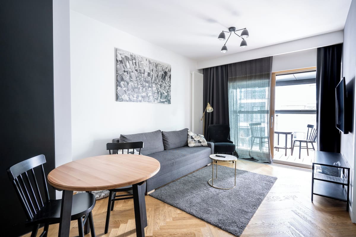A modern living room with a grey sofa, wooden dining table, and chairs. There is a black armchair, a small round table, and a TV on the wall.