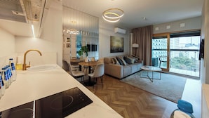 A modern kitchen with a built-in stove, a dining area with a round table and chairs, a sofa with cushions, and a large window offering a city view. - Vip Warsaw Apartments (Warsaw)