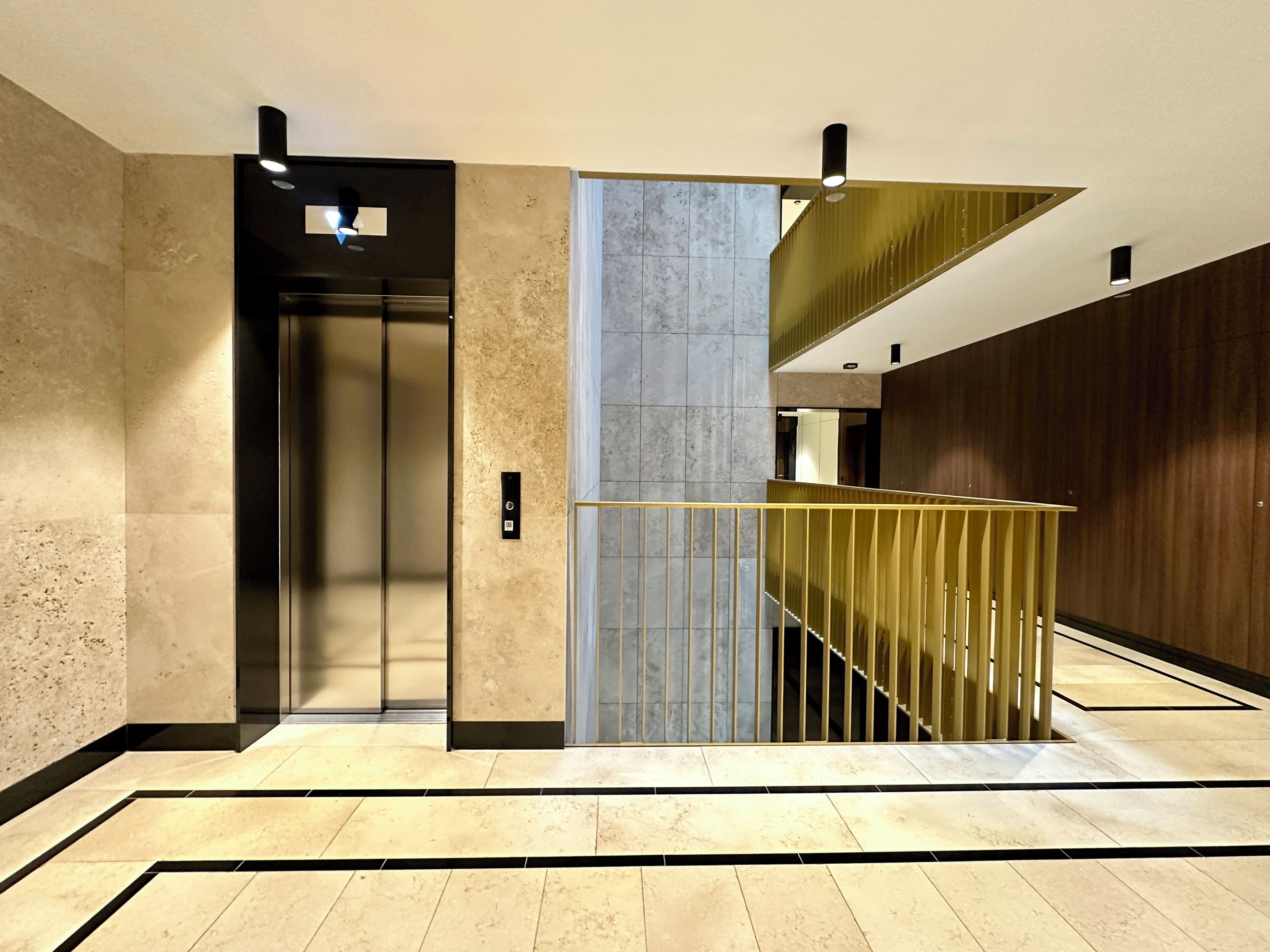A modern elevator with a metallic finish, a marble wall, and a wooden panel on the right.