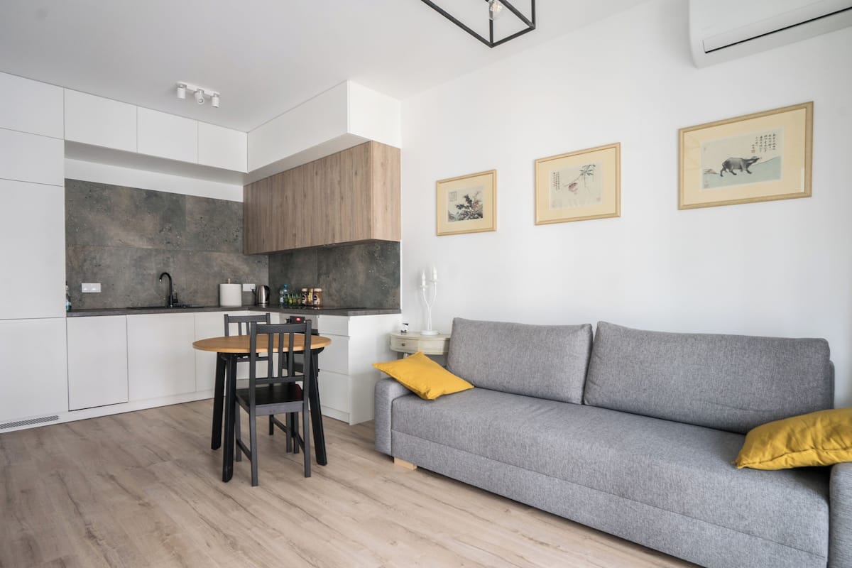 A modern kitchen with white cabinets, a wooden countertop, and a dining area with a table and chairs. There is a gray sofa with a yellow pillow against the wall.