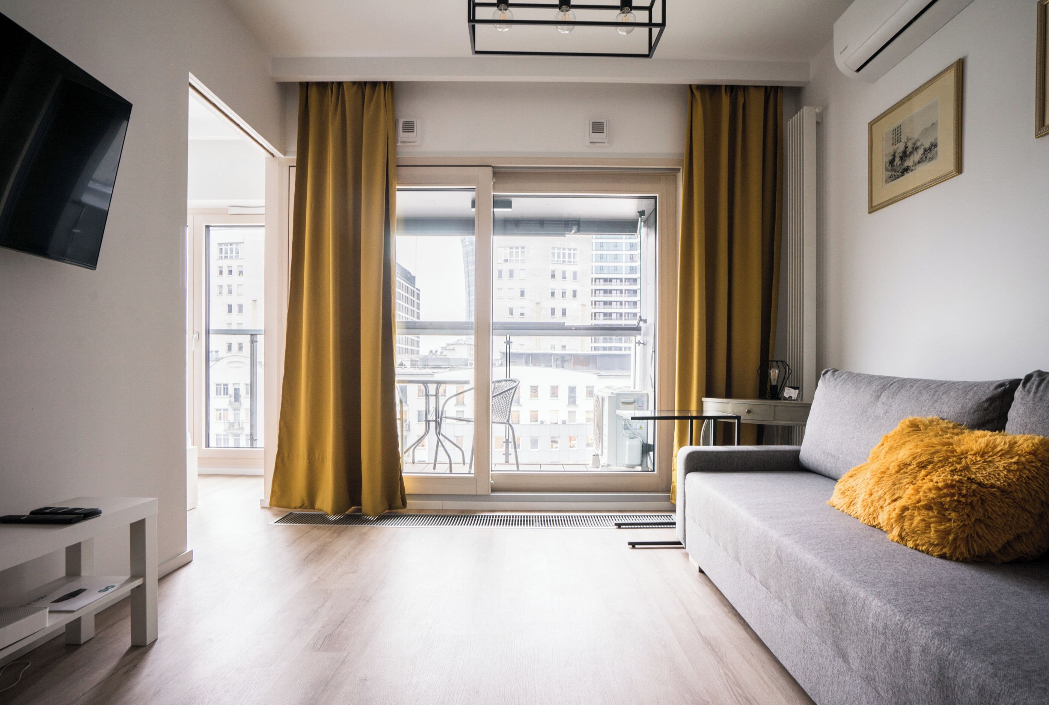 A modern living room with a grey sofa, a yellow throw pillow, and a large window with curtains.