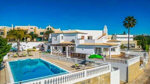 A poolside view of a resort with a swimming pool, lounge chairs, and a white building with a tiled roof. - Casa Oliveira - Four Bedroom Villa, Sleeps 8 (Albufeira)
