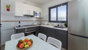 Een moderne keuken met witte kastjes, een roestvrijstalen koelkast, een magnetron en een spoelbak. Er staat een eettafel met stoelen en een schaal fruit.