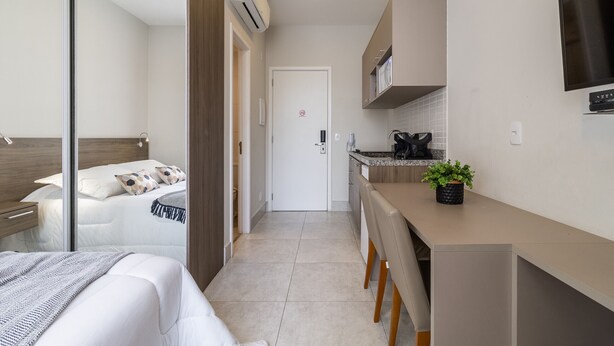 A compact hotel room with a bed, a desk, a kitchenette, and a TV mounted on the wall.