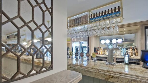 A bar area with a marble countertop, a variety of bottles and glasses, and a modern interior design. - The Queen Hotel Wetherspoon (Aldershot)