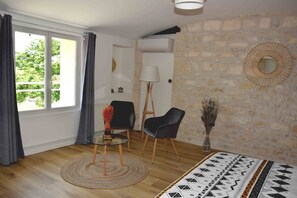 A modern living room with a stone wall, a round mirror, a black chair, a glass table with a vase, and a view through a large window.
