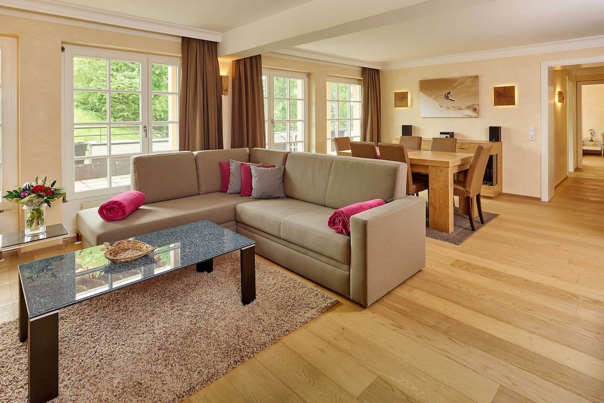 A modern living room with a beige sectional sofa, a glass-top coffee table, and a dining area with a wooden table and chairs.