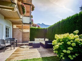 Un terrazzo in legno con mobili da esterno, un siepe e una montagna sullo sfondo.