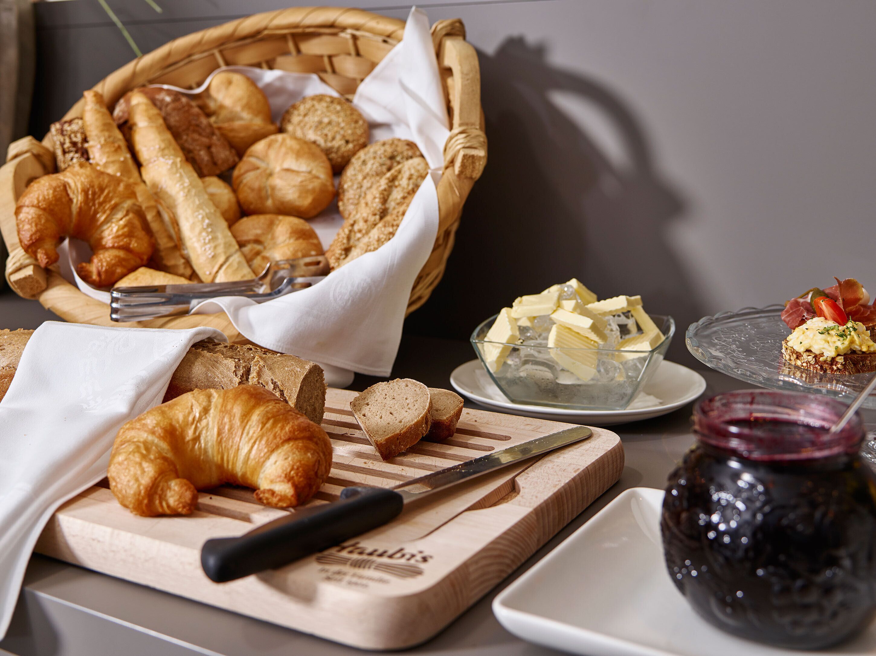 Ein Frühstückstisch mit Brot, Croissants, Butter, Marmelade und einem Messer auf einem Schneidebrett.