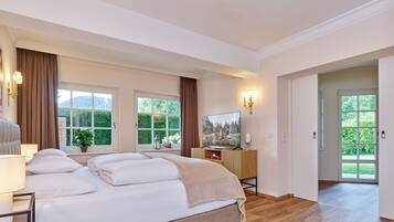 A spacious bedroom with a large bed, a television, and a view of greenery through the windows.