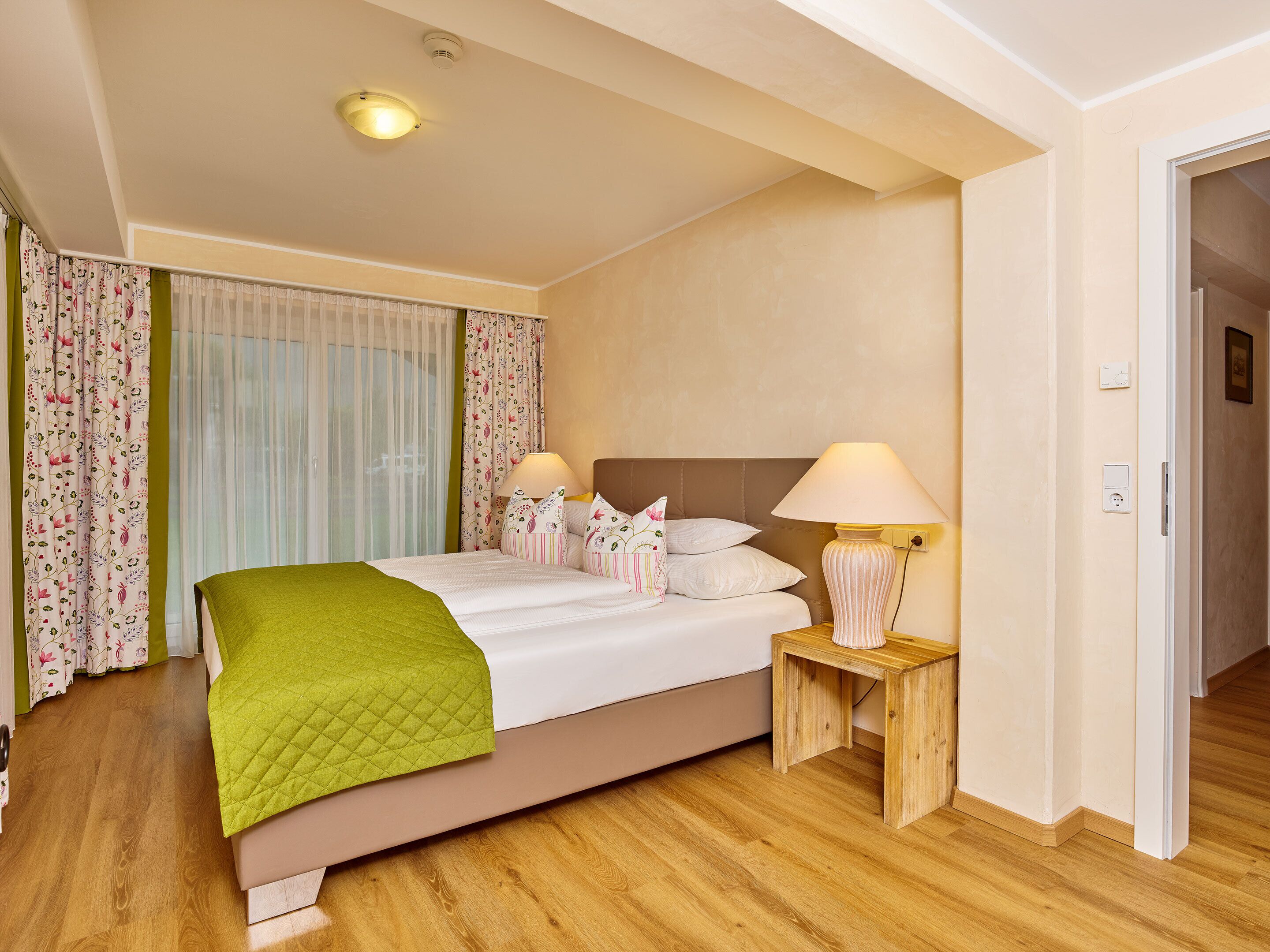 Een hotelkamer met een bed, twee nachtkastjes, een lamp, een raam met gordijnen en een deur.