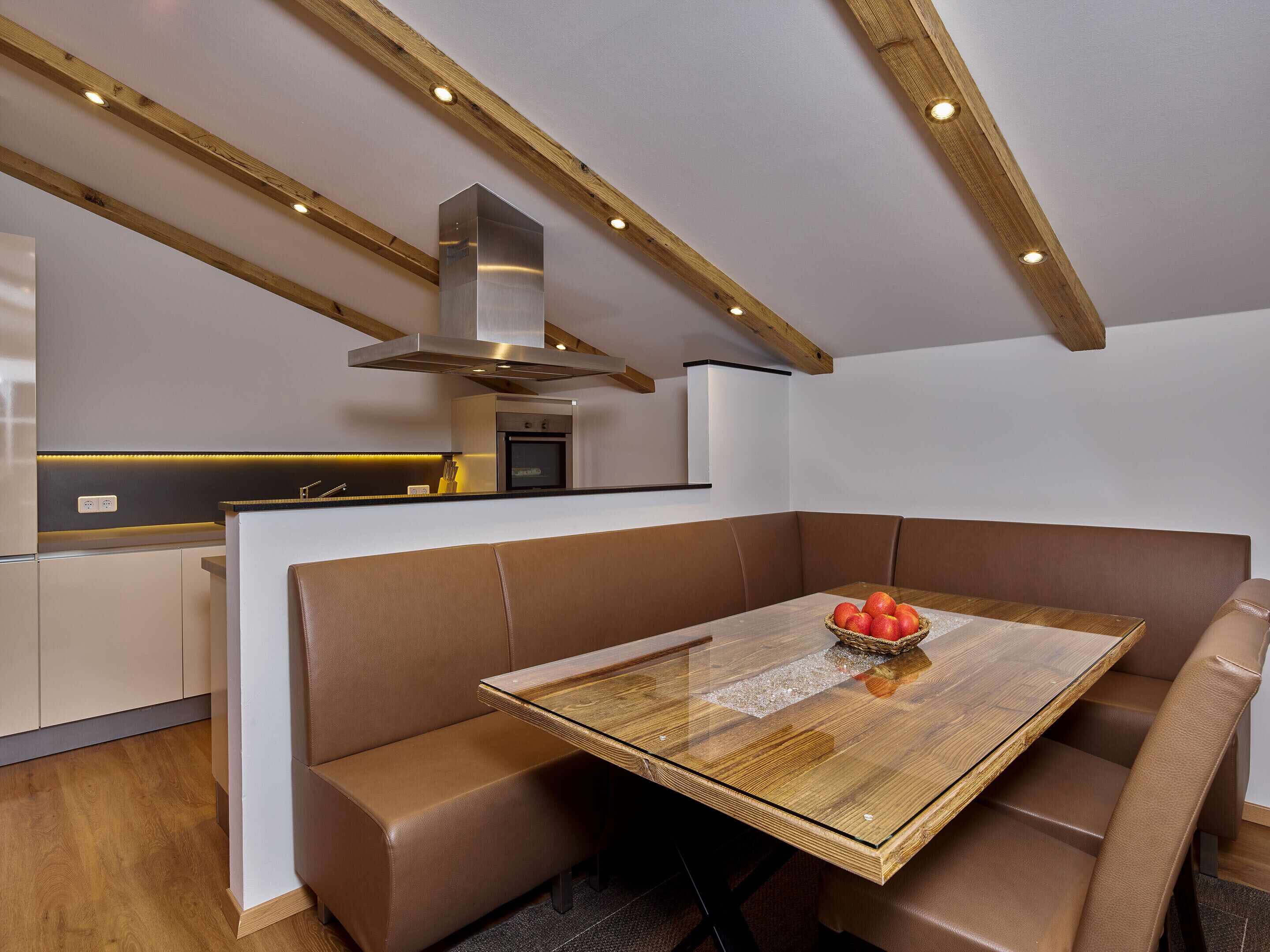 Una cocina moderna con vigas de madera, horno empotrado y comedor con mesa de vidrio y asientos de cuero.