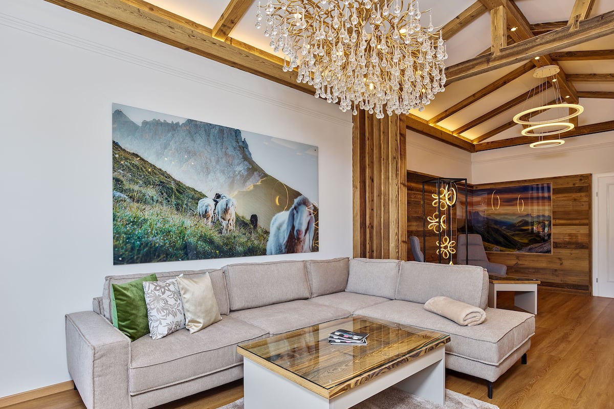 A modern living room with a large sectional sofa, a glass coffee table, a chandelier, and a large landscape painting on the wall.