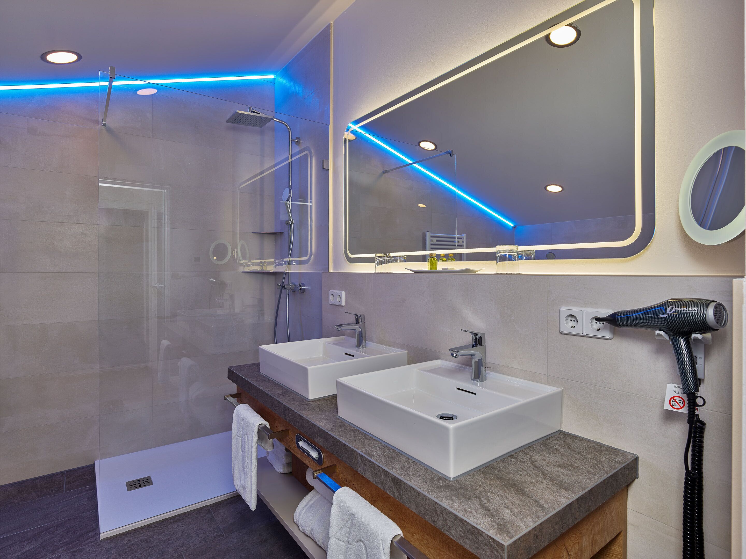 A modern bathroom with two sinks, a shower, and a hairdryer on a wall-mounted station.