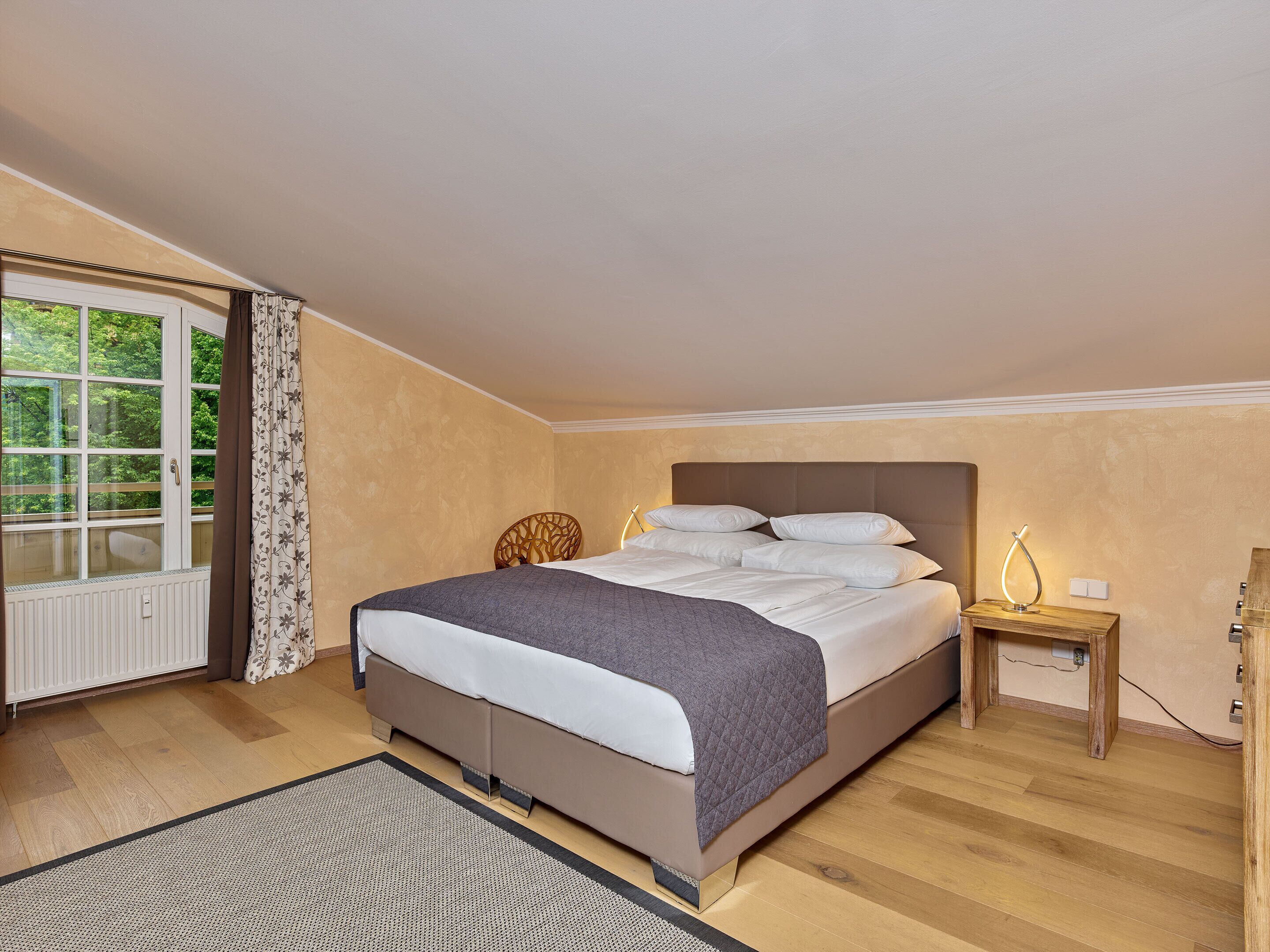 A bedroom with a bed, a nightstand, a window with curtains, and a view of greenery outside.