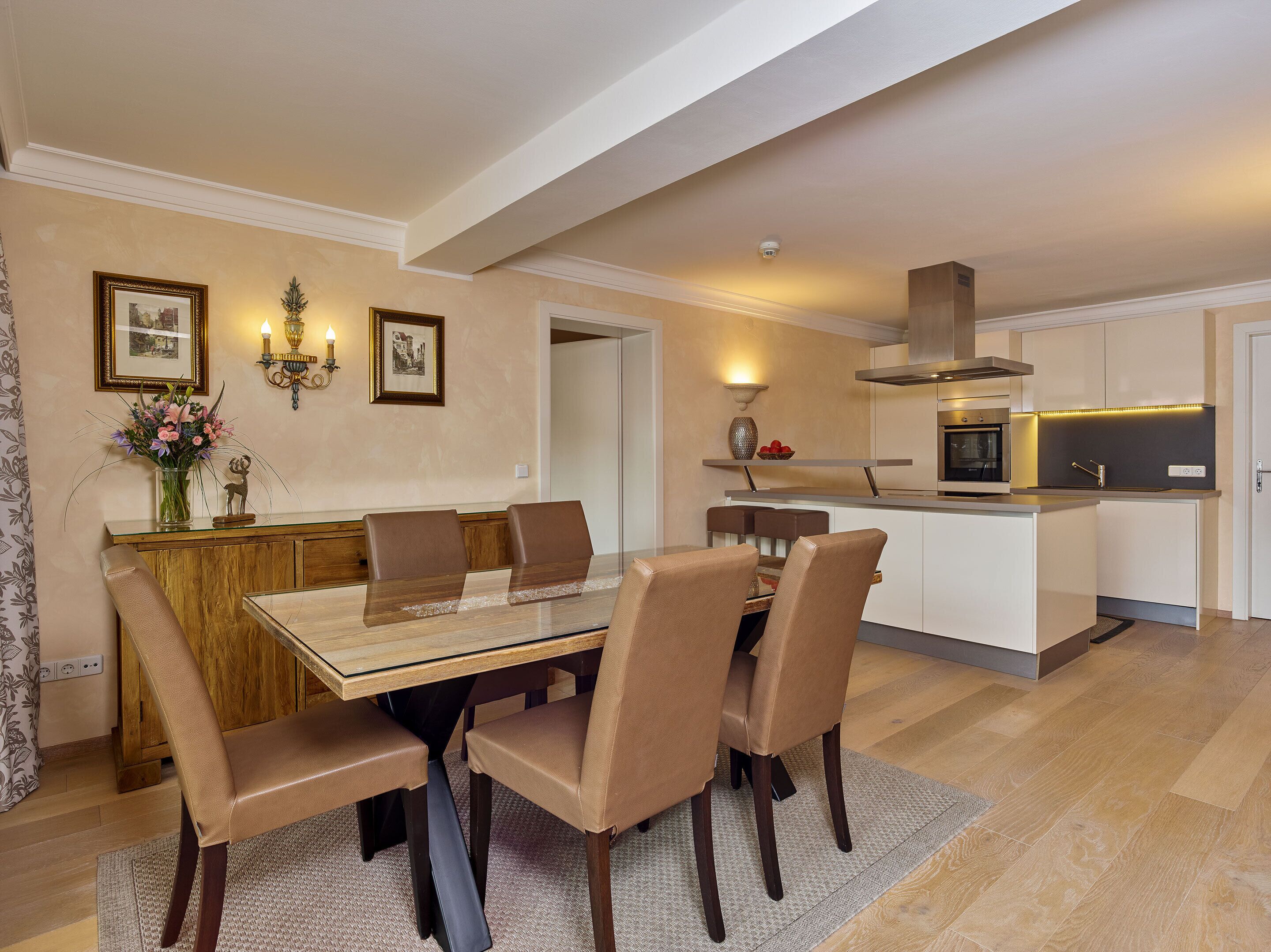 A modern dining area with a glass-top table, brown leather chairs, and a wooden cabinet with a vase of flowers.
