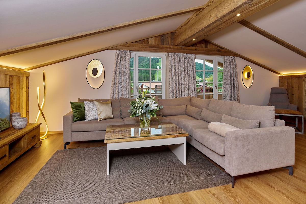 A cozy loft-style living room with a large sectional sofa, a glass coffee table, and a wooden ceiling.