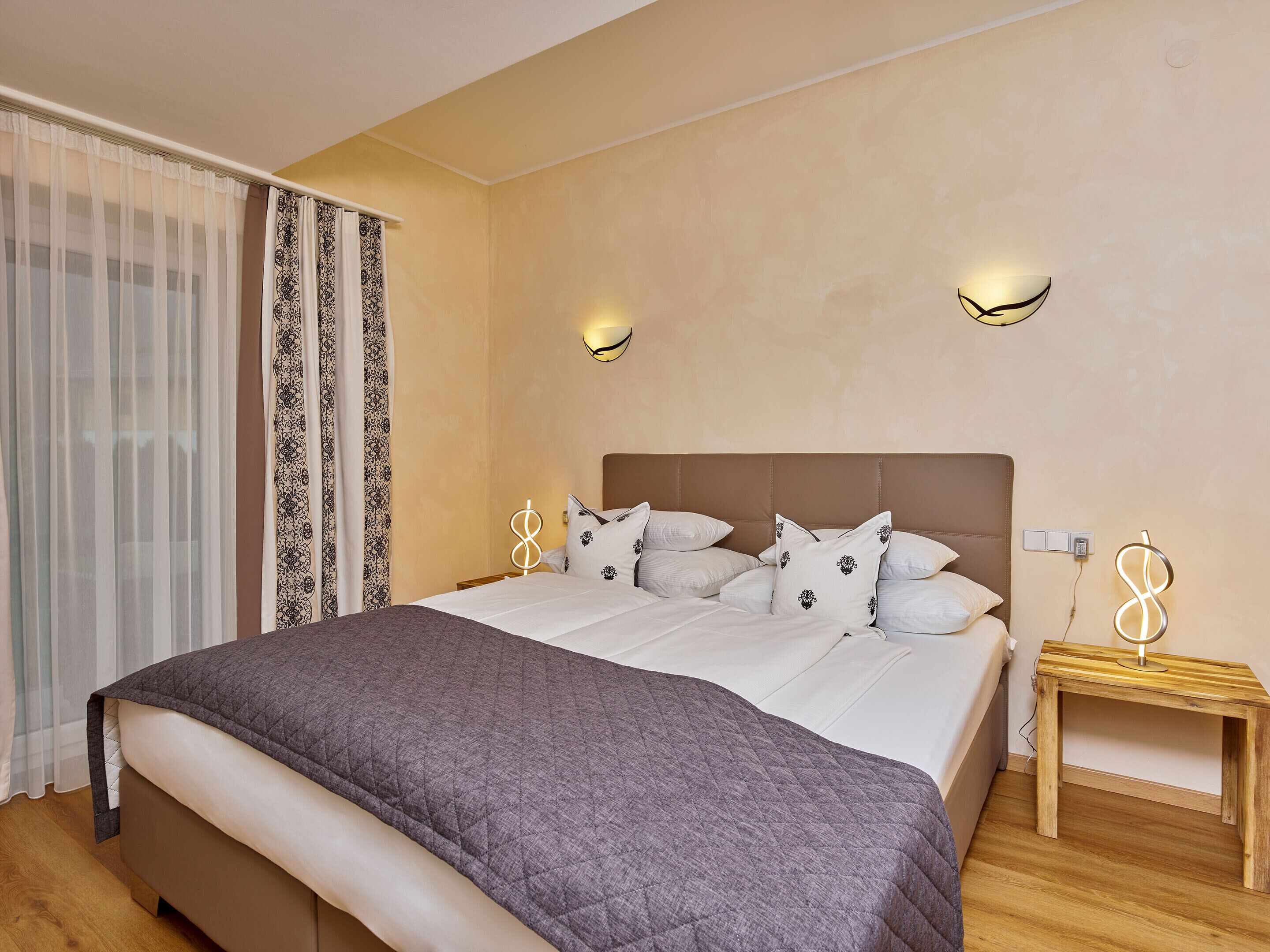 A bedroom with a large bed, two bedside tables, a wooden nightstand, and a wall-mounted light fixture.