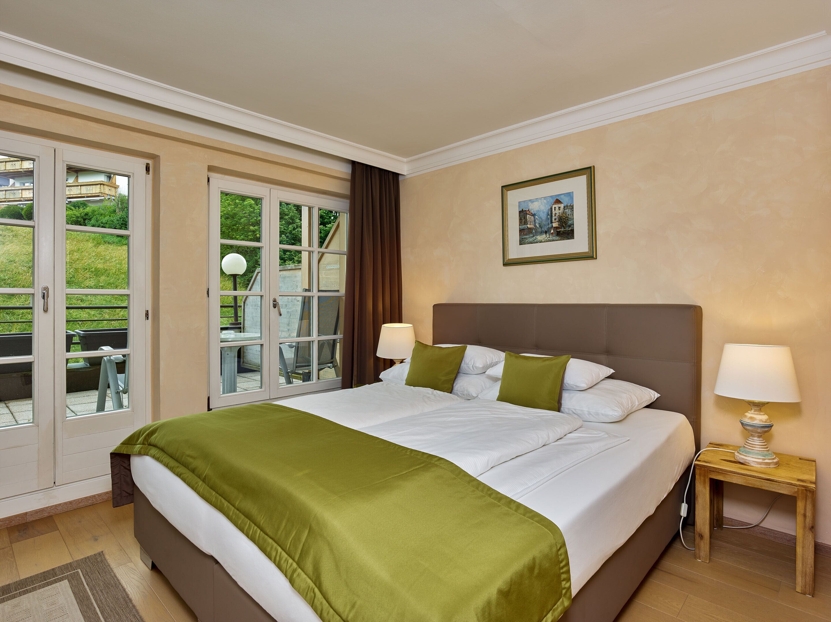 Habitación de hotel con una cama grande, dos lámparas de noche, una mesita con lámpara, un cuadro enmarcado en la pared y un ventanal corredero que da a un espacio exterior con mesa y sillas.