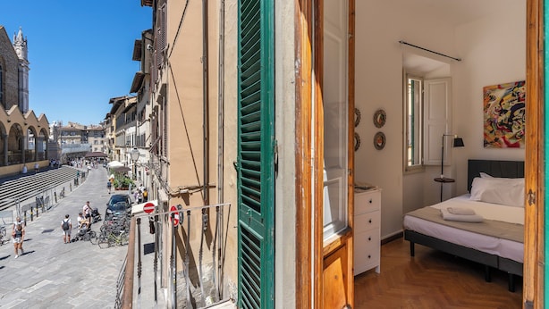 Una habitación con cama, cómoda y vista a la calle con edificios y gente.