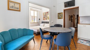 Una sala moderna con un sofá azul, una mesa de comedor redonda con sillas azules, una cama con ropa blanca y un televisor montado en la pared.