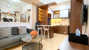A modern living room with a glass coffee table, a sofa with patterned cushions, and a kitchen area with wooden cabinets and a bar stool. - Great Choice And Modern 2Br At Transpark Juanda Bekasi Timur Apartment (Bekasi)