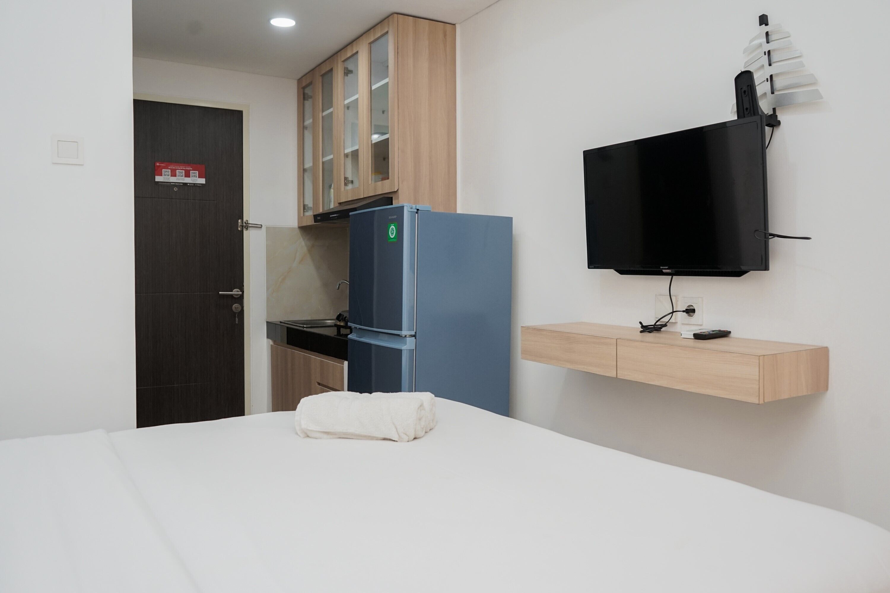 A compact hotel room with a bed, a blue refrigerator, a wooden cabinet, a mounted TV, and a small wooden shelf.