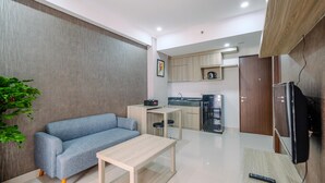 A modern living room with a sofa, a coffee table, and a TV. A kitchen area is visible in the background with wooden cabinets and a refrigerator. - Elegant And Comfy 2Br At Transpark Cibubur Apartment (Depok)