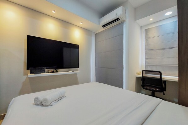 A modern hotel room with a flat-screen TV mounted on the wall, a bed with white linens, an air conditioning unit, and a window with blinds. - Modern Studio With Cozy Design Serpong Garden Apartment (Cisauk)