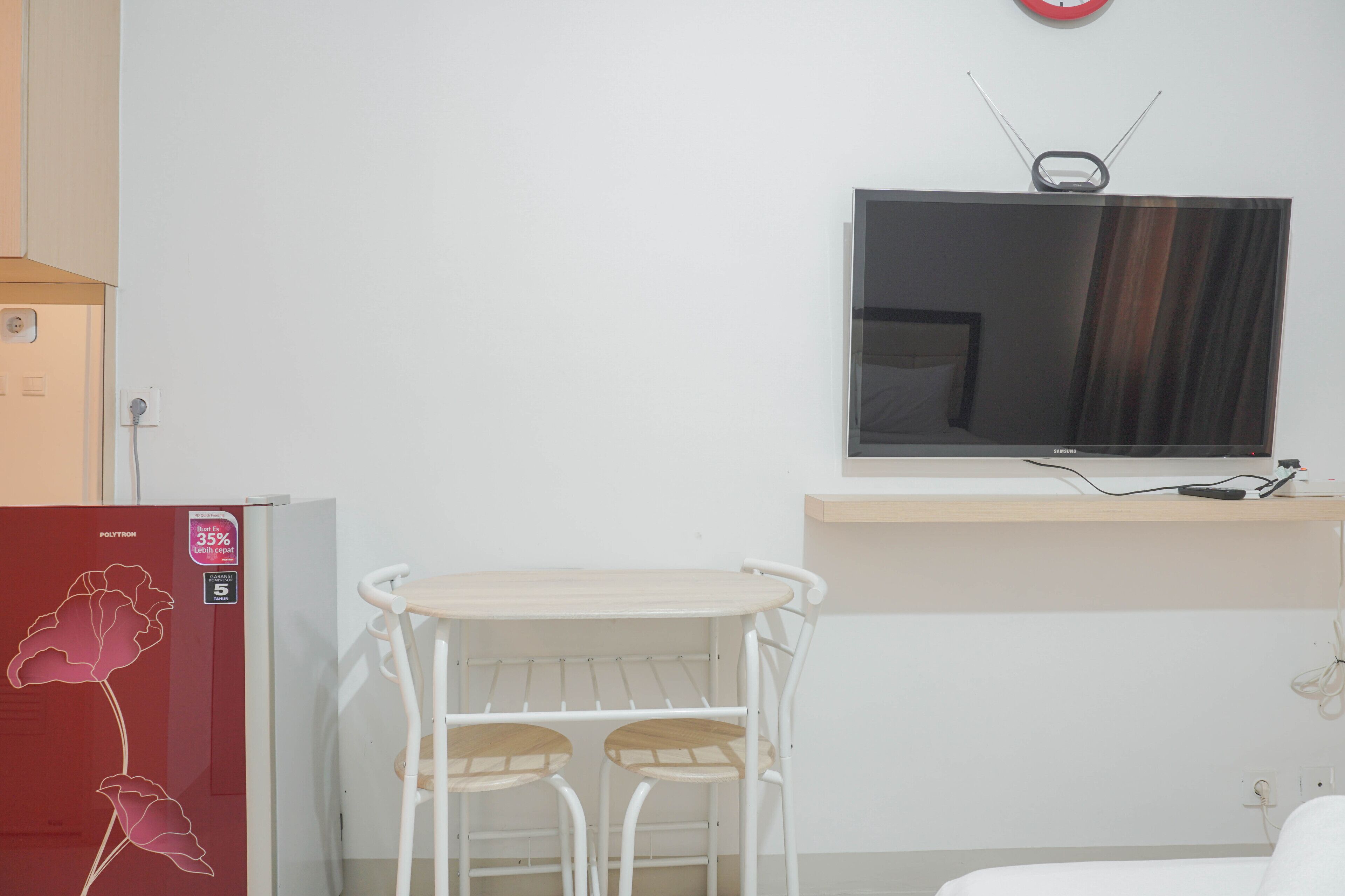 Ein kleines Zimmer mit Küchenzeile, einem kleinen Esstisch mit Stühlen, einem an der Wand montierten Fernseher und einem roten Kühlschrank mit Blumenmotiv.