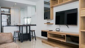A modern kitchen with a built-in stove, a refrigerator, and a large TV mounted on the wall. There is a wooden dining table with stools and a small cabinet below the TV.
