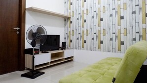 Ein Zimmer mit Fernseher, Standventilator, grünem Sofa und einer Wand mit Muster.