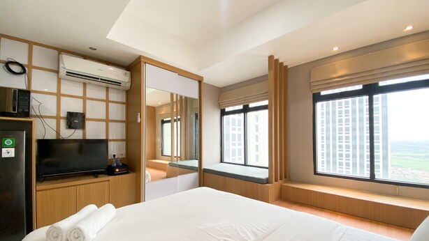 A hotel room with a large bed, a television, an air conditioning unit, and a window with a city view. - Nice Studio Apartment At Pollux Chadstone (Cikarang)