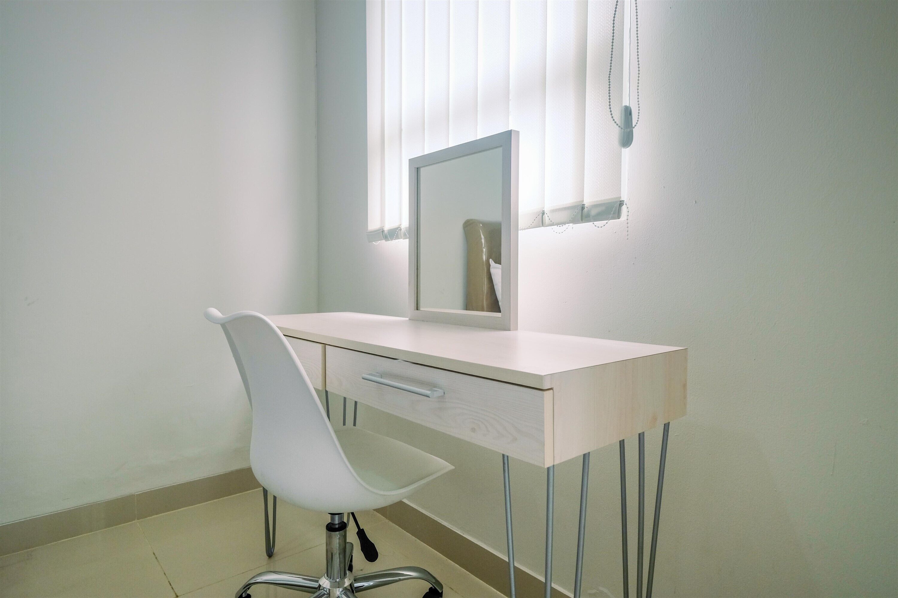 A minimalist office desk with a white chair, a mirror, and a window with blinds.