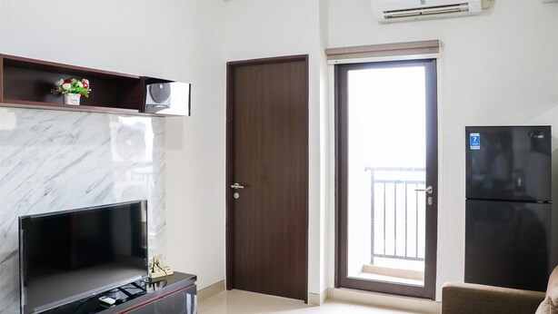 A modern living room with a flat-screen TV, a dark wooden door, a balcony with a view, and a black refrigerator. - Comfort And Cozy Stay 2Br At 29Th Floor Transpark Cibubur Apartment (Depok)