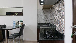 A compact kitchen with a black countertop, a sink, and a stove. There is a dining table with chairs and a small shelf with decorative items. - Comfort And Cozy Stay 2Br At 29Th Floor Transpark Cibubur Apartment (Depok)