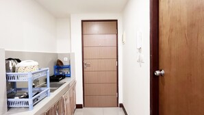 A narrow hallway with a wooden door, a small kitchen area with blue baskets and dish racks, and a white wall with a power outlet. - Compact Studio Room Apartment Vida View Makassar (Makassar)