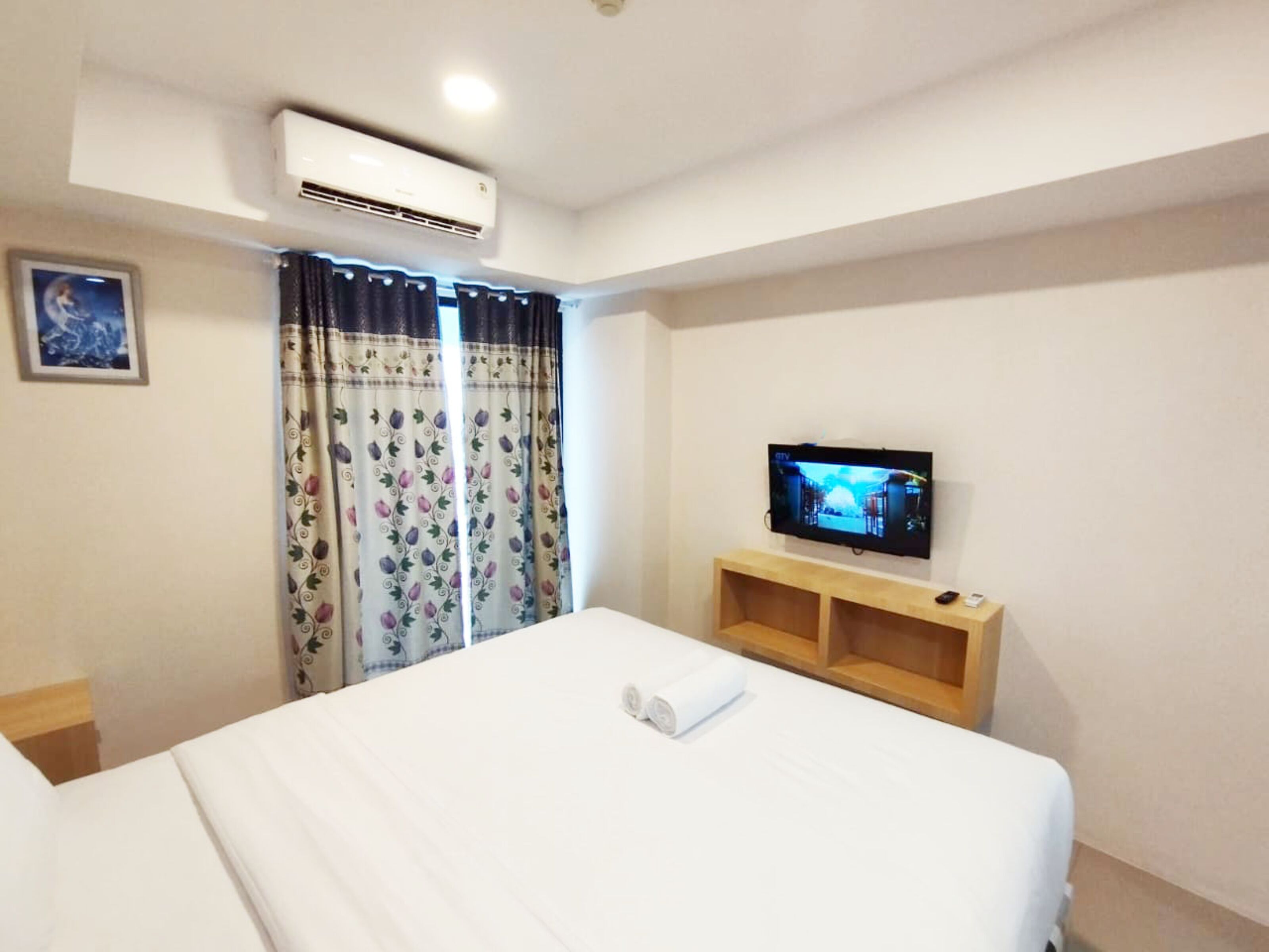 A hotel room with a bed, an air conditioning unit, a television on a wooden stand, and a window with curtains.