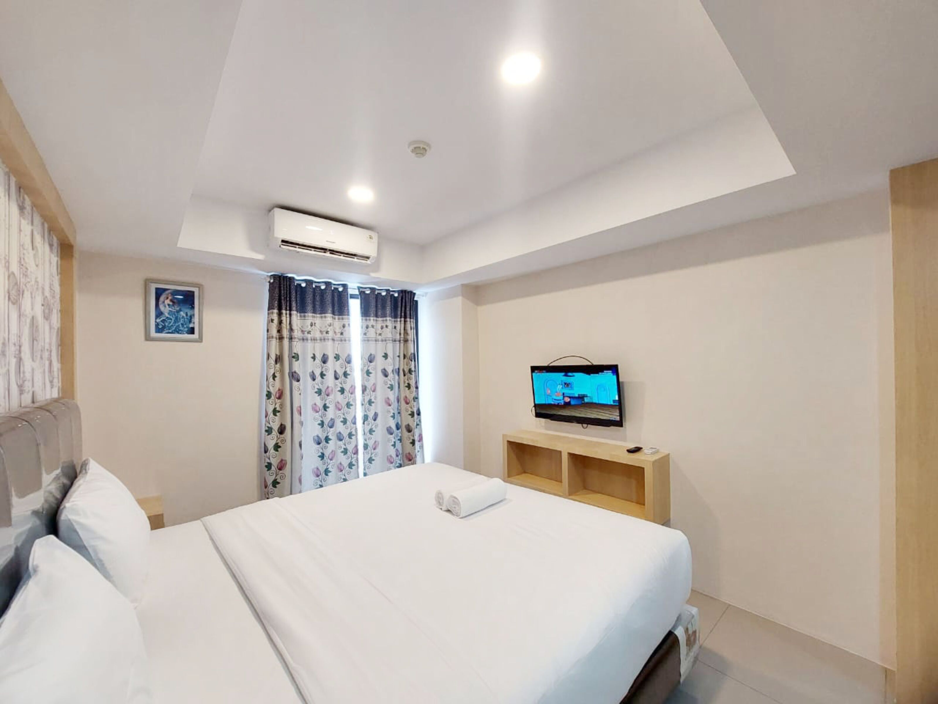 A modern hotel room with a large bed, a flat-screen TV mounted on the wall, and a ceiling-mounted air conditioning unit.