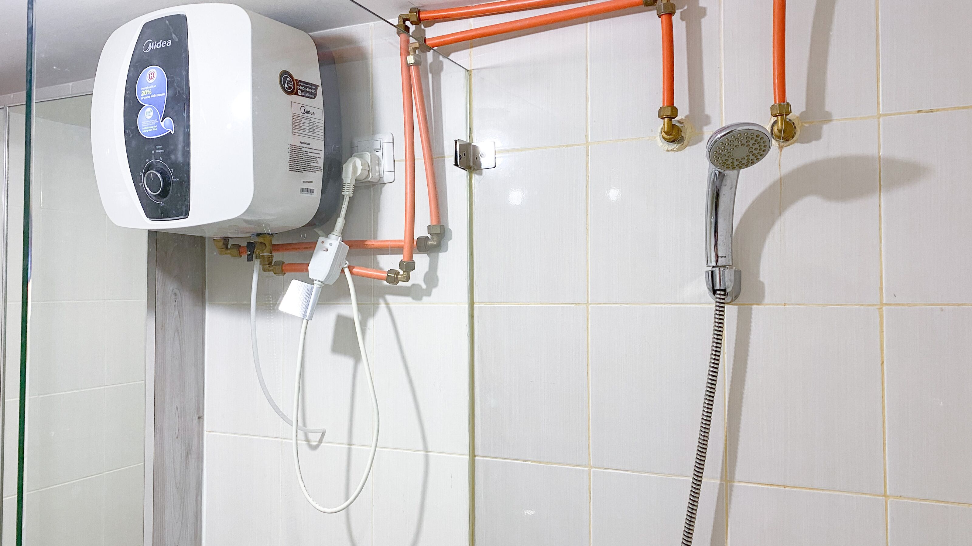 A bathroom with a white tiled wall, a white shower unit with orange pipes, and a handheld showerhead.