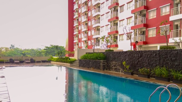 A swimming pool with a stone wall and metal railings, surrounded by greenery and a red building with balconies. - Homey Studio Room At Taman Melati Margonda Apartment (Depok)