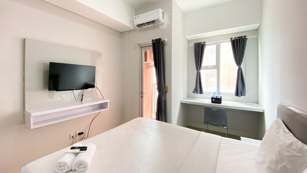 A hotel room with a bed, a TV mounted on the wall, a window with curtains, and an air conditioning unit. - Nice And Cozy Studio At Transpark Juanda Bekasi Timur Apartment (Bekasi)