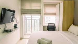 A hotel room with a bed, a TV, a desk, and a window with blinds. - Studio Room With Cozy Style At Serpong Garden Apartment (Cisauk)