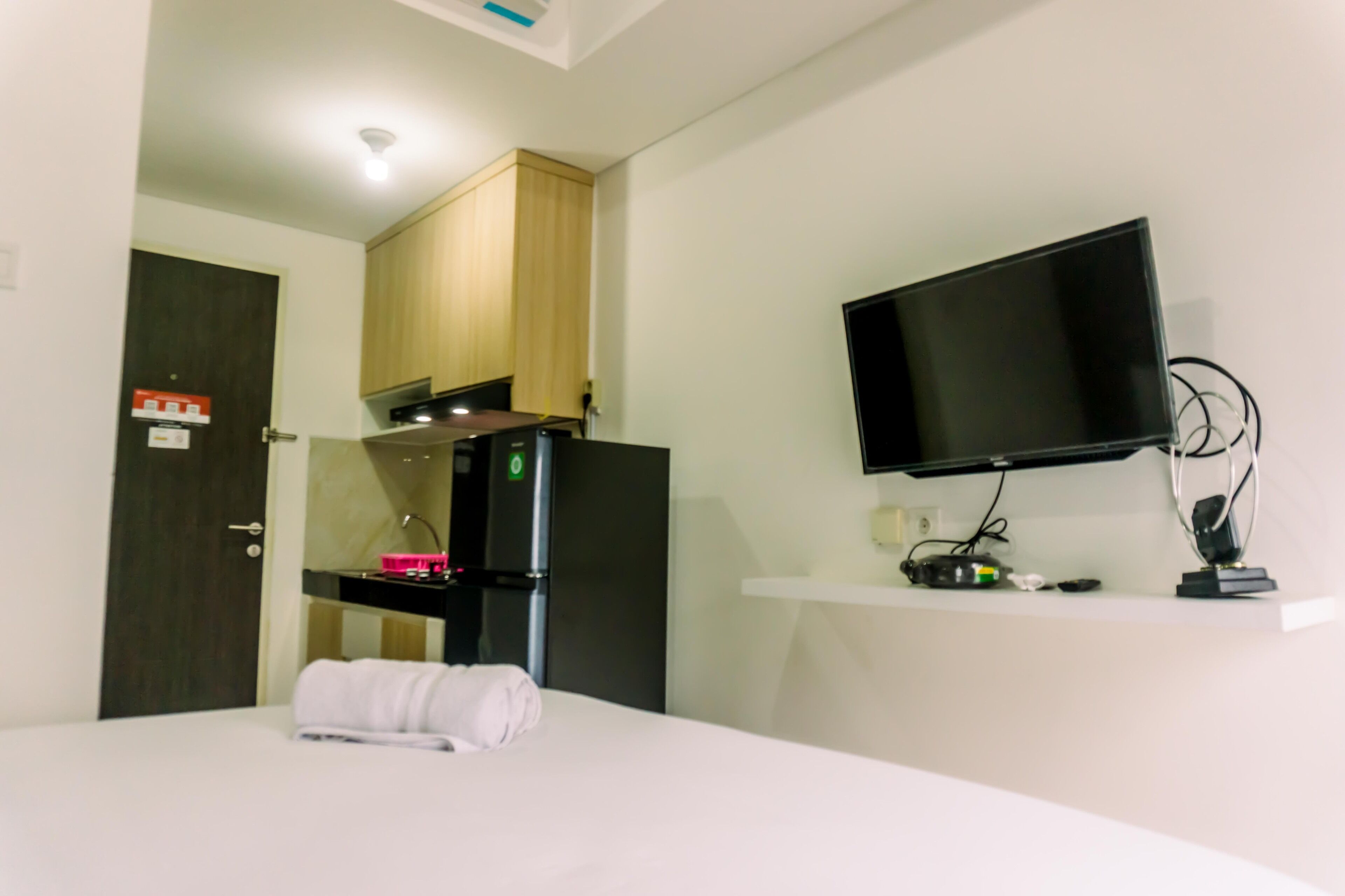 A hotel room with a bed, a flat-screen TV mounted on the wall, a small kitchenette with a refrigerator, and a wooden door.