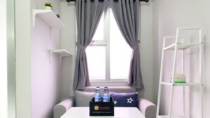 A room with a grey sofa, a small table with a tray of water bottles and a tray bag, a window with grey curtains, and a white shelf with a plant.